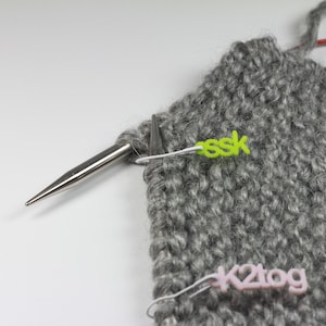 May include: Close-up of a grey knitted fabric with two stitch markers. One marker is green and says "ssk" and the other is pink and says "k2tog".