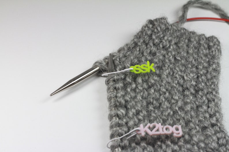May include: Close-up of a grey knitted fabric with two stitch markers. One marker is green and says "ssk" and the other is pink and says "k2tog".