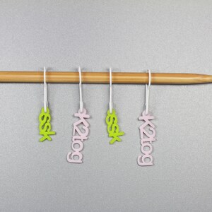 May include: Four stitch markers hanging on a wooden knitting needle. Two markers are pink with the text "k2tog" and two are green with the text "ssk".