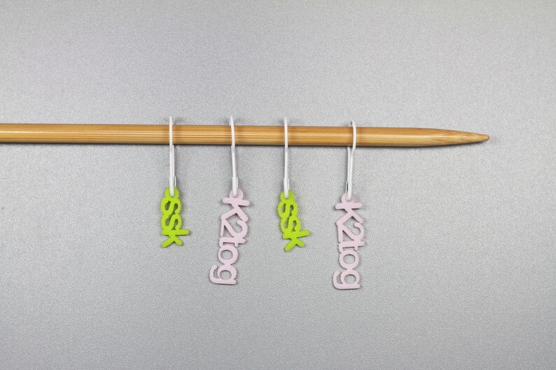 May include: Four stitch markers hanging on a wooden knitting needle. Two markers are pink with the text "k2tog" and two are green with the text "ssk".
