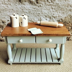 May include: A miniature wooden kitchen island with a light blue base, two drawers with white knobs, and a brown countertop. On the surface are two small white canisters with black speckles, a wooden rolling pin, and a folded piece of paper.