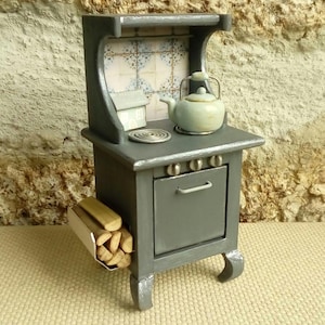 May include: A miniature gray wooden stove featuring a light blue kettle on the stovetop, a tiled backsplash with blue and white patterns, three control knobs, an oven door with a handle, and a side rack filled with small wooden logs.