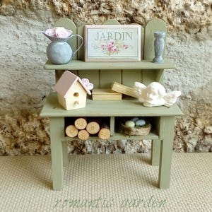 Könnte beinhalten: Ein Miniatur-Garten-Anrichte in Salbeigrün mit zweistufigem Design. Es verfügt über ein kleines Vogelhaus, ein Schild mit der Aufschrift "JARDIN", einen dekorativen Engel und einen kleinen grauen Krug mit rosa Rosen. Die Bank hat ein unteres Regal mit Holzscheiten und einem Nest.