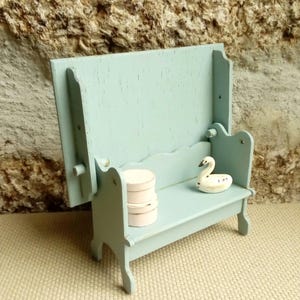 May include: A light blue wooden bench with a high backrest and scalloped edges. On the seat, there are two small stacked round white containers and a small white swan figurine with black details. The bench has a distressed paint finish.