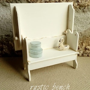 May include: A small white wooden bench with a high backrest and side arms. On the seat, there are two stacked light blue circular containers and a small white swan figurine with a decorative neck detail. The bench features a distressed finish.