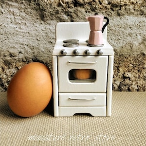 May include: A miniature white wooden stove with four grey burners and a silver handle on the oven door. A small pink moka pot with a black handle sits on the stovetop. A large brown egg is positioned to the left of the miniature stove.