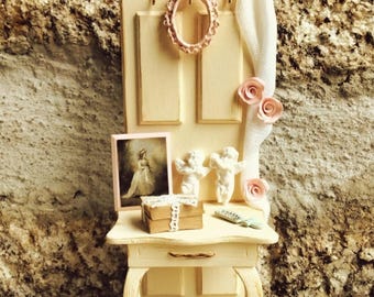 May include: A miniature cream-colored wooden vanity with a tall back panel, featuring a small pink oval frame, a white fabric drape with three pink roses, two white cherub figurines, a framed portrait, a stack of books, and a small hairbrush.