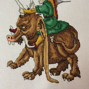 May include: Cross-stitch embroidery of a person in a green robe and feathered hat riding a brown, furry, mythical creature with a long snout and claws.