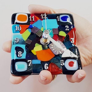 May include: A square clock with a mosaic design featuring colorful glass pieces. The clock face is black with white numbers and hands. The clock is held in a hand.