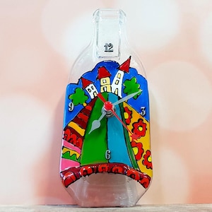 May include: A colorful glass bottle clock with a whimsical painted design of houses on a hill. The clock face features a blue background with white numbers and a silver hand.