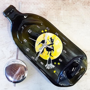 May include: A green glass bottle clock with a yellow moon and a black silhouette of a witch flying on a broomstick. The clock face has black numbers and silver hands.