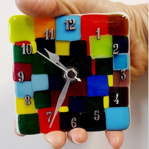 Puede incluir: Un reloj cuadrado colorido con un diseño de mosaico. La esfera del reloj está hecha de pequeños cuadrados de vidrio en varios colores, incluyendo rojo, amarillo, azul, verde y negro. Los números en la esfera del reloj son plateados y las manecillas son plateadas.