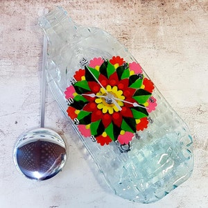 May include: A clear glass bottle clock with a colorful floral design. The clock face is painted with red, green, yellow, and pink flowers. The clock has a pendulum and is hanging on a wall.