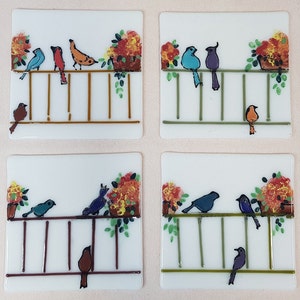 May include: Four square glass coasters with a white background and a painted design of birds on a railing with flowers in pots. The birds are blue, red, orange, and purple.