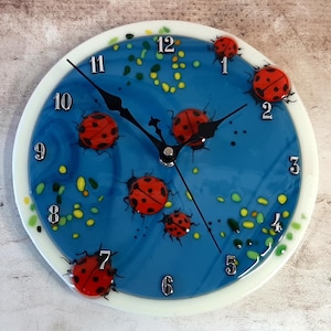 May include: A blue glass clock face with black Roman numeral hour markers and black hands. The clock face is decorated with red ladybugs and yellow and green dots.