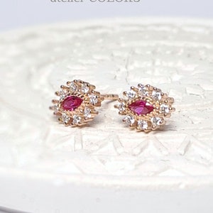 Op de afbeelding: Een paar gouden oorbellen met een rode edelsteen omringd door kleinere witte edelstenen.