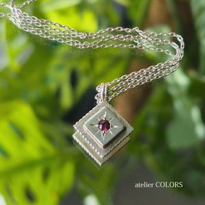 May include: A silver chain necklace with a square pendant featuring a red gemstone and a star design. The pendant is layered with a second square frame. The necklace is photographed against a green leafy background.