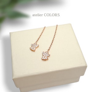 Puede incluir: Un par de pendientes colgantes de oro rosa con un peque&ntilde;o dise&ntilde;o brillante que parece un diamante. Los pendientes se muestran en una caja blanca con el texto "atelier COLORS" en la parte superior.