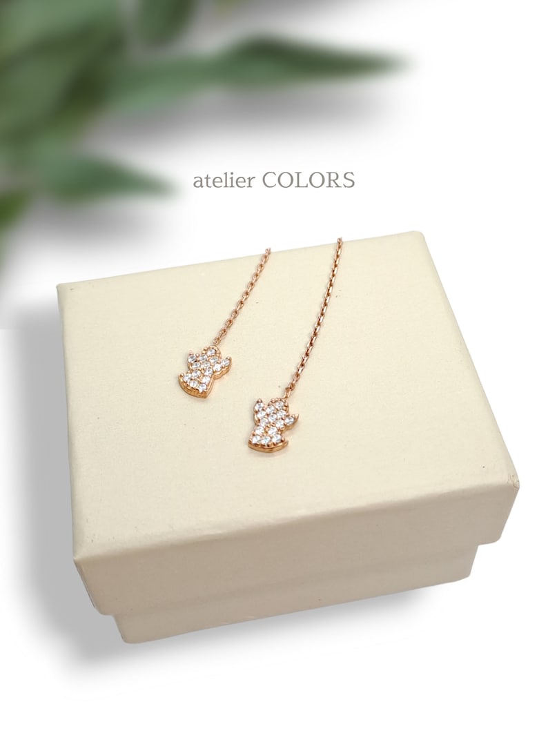 Puede incluir: Un par de pendientes colgantes de oro rosa con un peque&ntilde;o dise&ntilde;o brillante que parece un diamante. Los pendientes se muestran en una caja blanca con el texto "atelier COLORS" en la parte superior.
