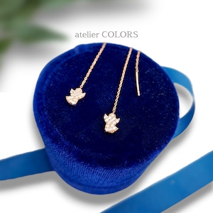Puede incluir: Un par de pendientes de oro en forma de &aacute;ngel con piedras de circonita c&uacute;bica transparentes, presentados en una caja de terciopelo azul con una cinta azul. La caja tiene el texto "atelier COLORS" en la parte superior.