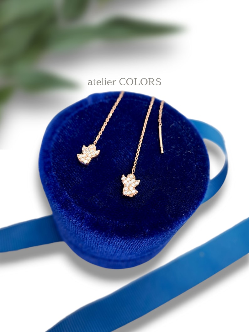 Puede incluir: Un par de pendientes de oro en forma de &aacute;ngel con piedras de circonita c&uacute;bica transparentes, presentados en una caja de terciopelo azul con una cinta azul. La caja tiene el texto "atelier COLORS" en la parte superior.