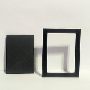May include: Two black rectangular objects on a white surface. One is a solid, closed rectangle with a textured surface. The other is a hollow, open rectangular frame.
