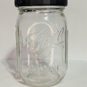 May include: A clear glass Ball Mason jar with a black lid. The jar has the word "Ball" and "MASON" embossed on the front. The jar is empty and ready to be filled. The jar is a classic design for preserving and storage.