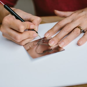 May include: A person is using a tool to etch a design into a copper sheet. The design is a heart with swirls around it.