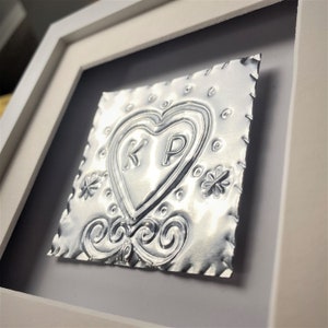 May include: A silver metal heart with the initials "KP" embossed inside, framed in a white frame. The heart is surrounded by decorative details, including swirls and flowers.