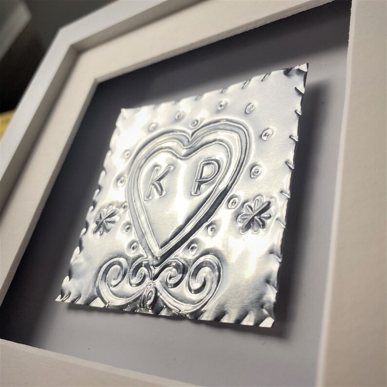May include: A silver metal heart with the initials "KP" embossed inside, framed in a white frame. The heart is surrounded by decorative details, including swirls and flowers.
