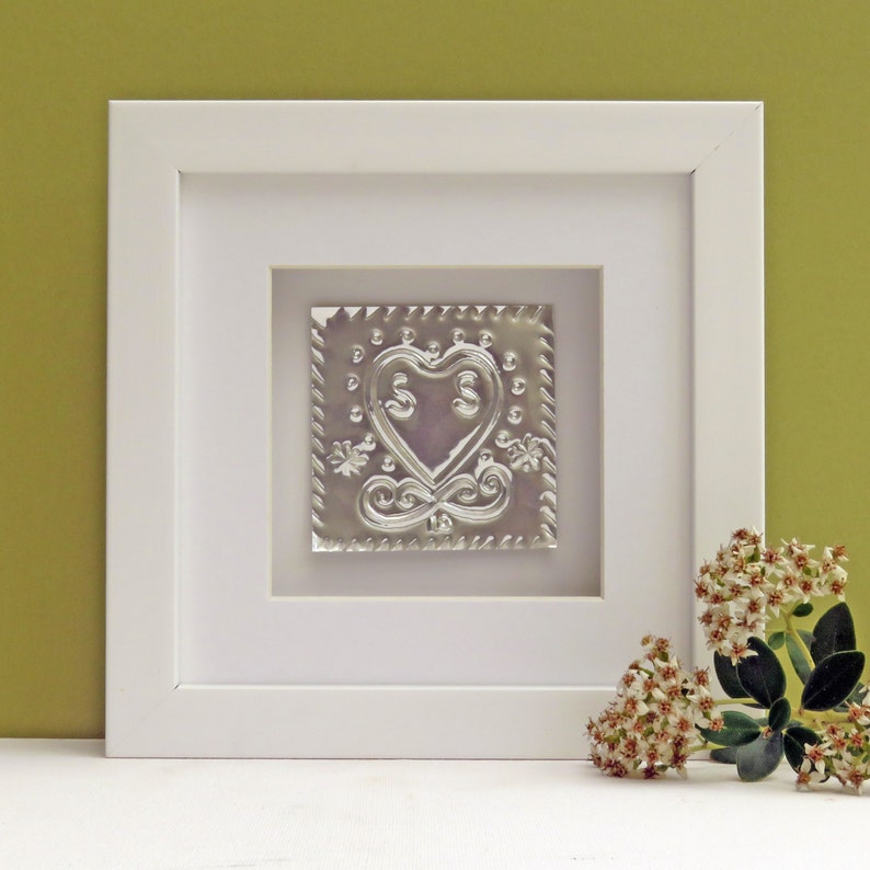 May include: A white framed picture with a silver heart design inside. The heart is embossed with two S's and has a decorative border. The picture is sitting on a white surface with a small bouquet of white flowers to the right.