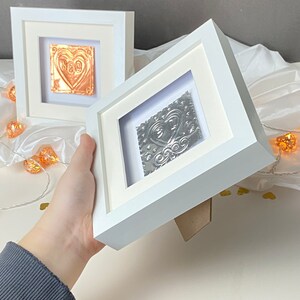 May include: Two white picture frames with a silver and copper heart design inside. The frames are on a white surface with string lights.