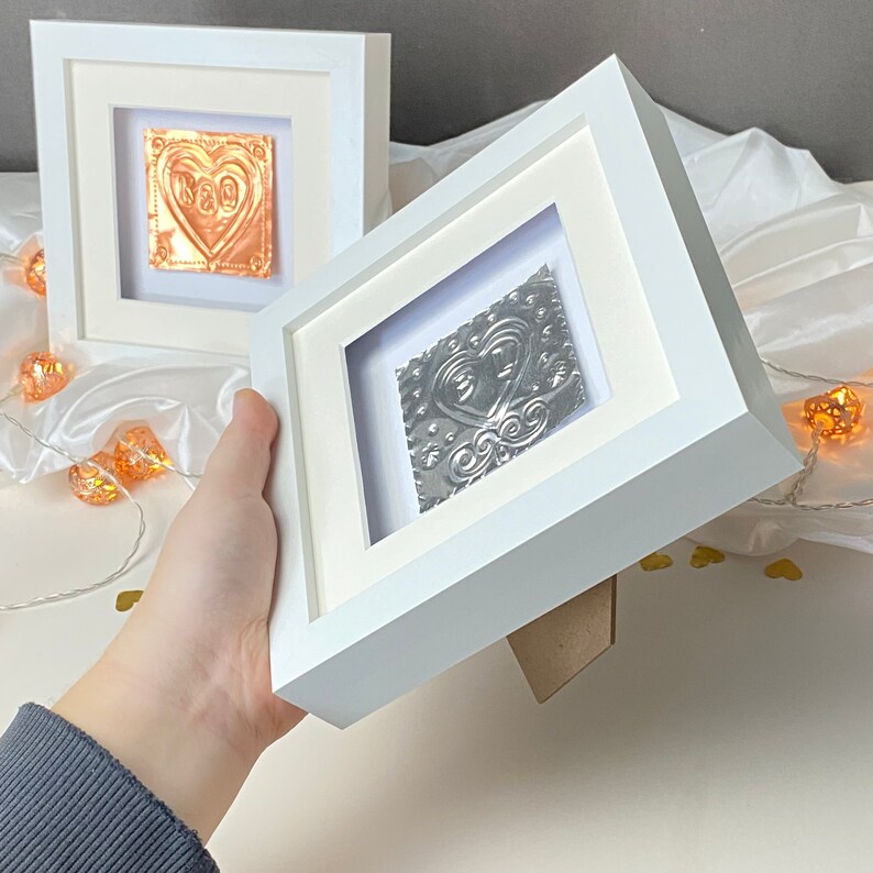 May include: Two white picture frames with a silver and copper heart design inside. The frames are on a white surface with string lights.