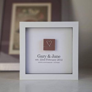 May include: A white frame with a brown square tile inside. The tile has a wire heart on it. The frame has the text "Gary & Jane est. 22nd February 2012 NINTH ANNIVERSARY - POTTERY" printed on it.