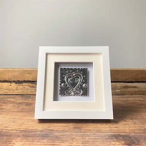 May include: A white framed picture with a silver metal heart with the initials "KP" inside. The heart is surrounded by a decorative border.