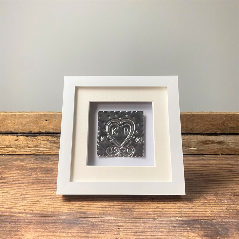 May include: A white framed picture with a silver metal heart with the initials "KP" inside. The heart is surrounded by a decorative border.