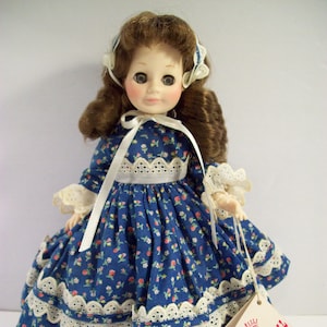 May include: A vintage porcelain doll wearing a blue and white floral dress with lace trim. The doll has brown hair and blue eyes. The doll is holding a tag that reads "A Royal Doll, Trademark for a product".