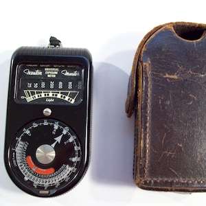 May include: A black Weston Universal Exposure Meter Master light meter with a leather case. The meter has a dial with numbers from 0 to 1500 and a needle that points to the correct exposure setting. The case is brown leather with a flap that closes with a snap.