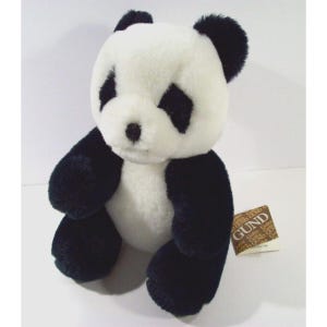 May include: A black and white plush panda bear sitting on a white background. The panda has black fur on its ears, legs, and around its eyes. The panda is holding a brown tag that says "GUND".