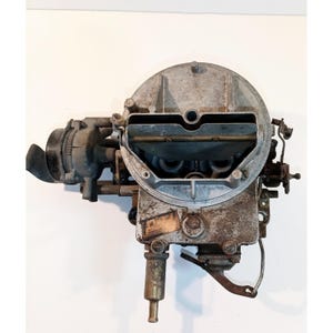 May include: A metallic automotive carburetor featuring a circular air intake housing, a rectangular base with a weathered finish, various mechanical linkages, a side-mounted black cylindrical component, and a small identification plate.