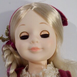 May include: A porcelain doll with blonde hair styled in a braid. She is wearing a burgundy dress with a white lace collar and a burgundy headband.