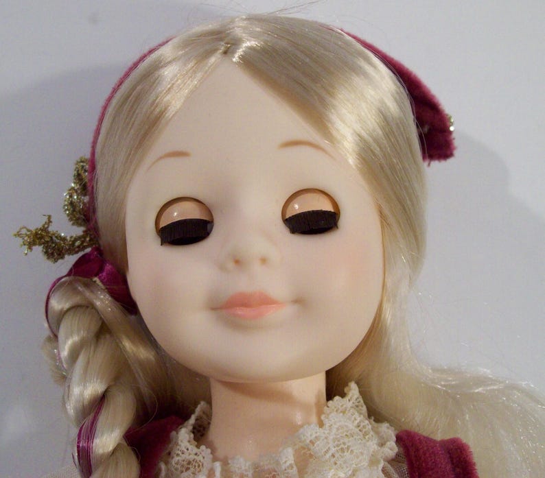 May include: A porcelain doll with blonde hair styled in a braid. She is wearing a burgundy dress with a white lace collar and a burgundy headband.