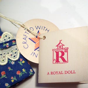 May include: A blue and white floral dress with a white lace trim. The dress has a tag that says "Crafted with Z" and another tag that says "A Royal Doll" with a red letter R in a castle.