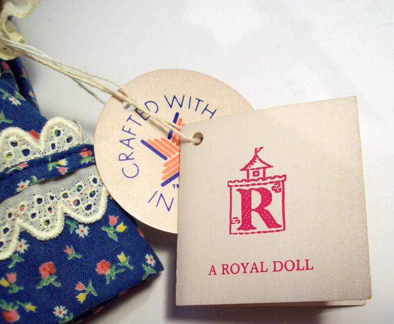 May include: A blue and white floral dress with a white lace trim. The dress has a tag that says "Crafted with Z" and another tag that says "A Royal Doll" with a red letter R in a castle.
