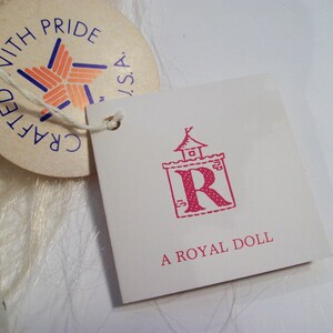 May include: A white tag with pink text that reads "A Royal Doll" and a pink illustration of a castle with a letter R inside. The tag is attached to a white fabric with a smaller tag that reads "Crafted with Pride, USA".