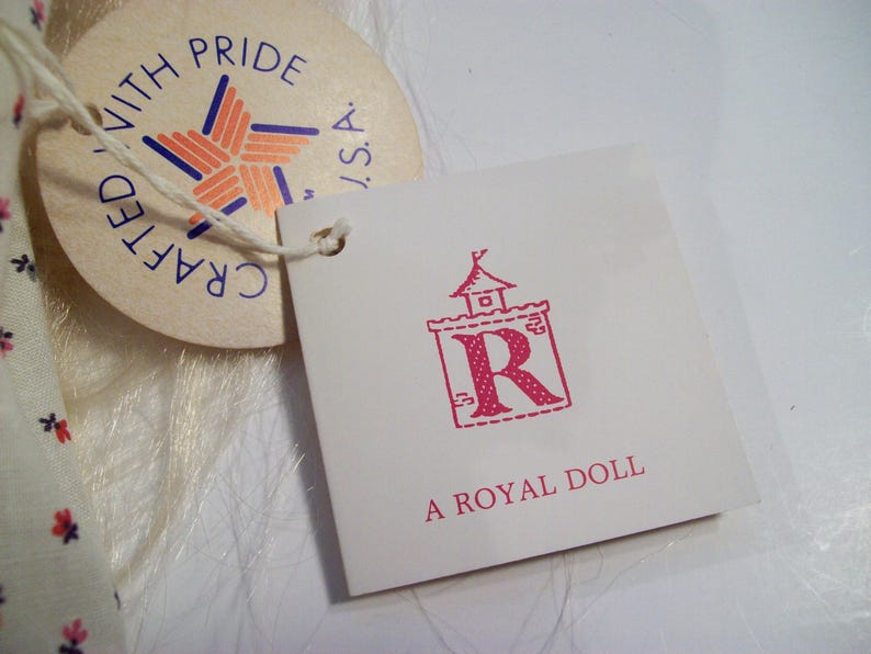 May include: A white tag with pink text that reads "A Royal Doll" and a pink illustration of a castle with a letter R inside. The tag is attached to a white fabric with a smaller tag that reads "Crafted with Pride, USA".