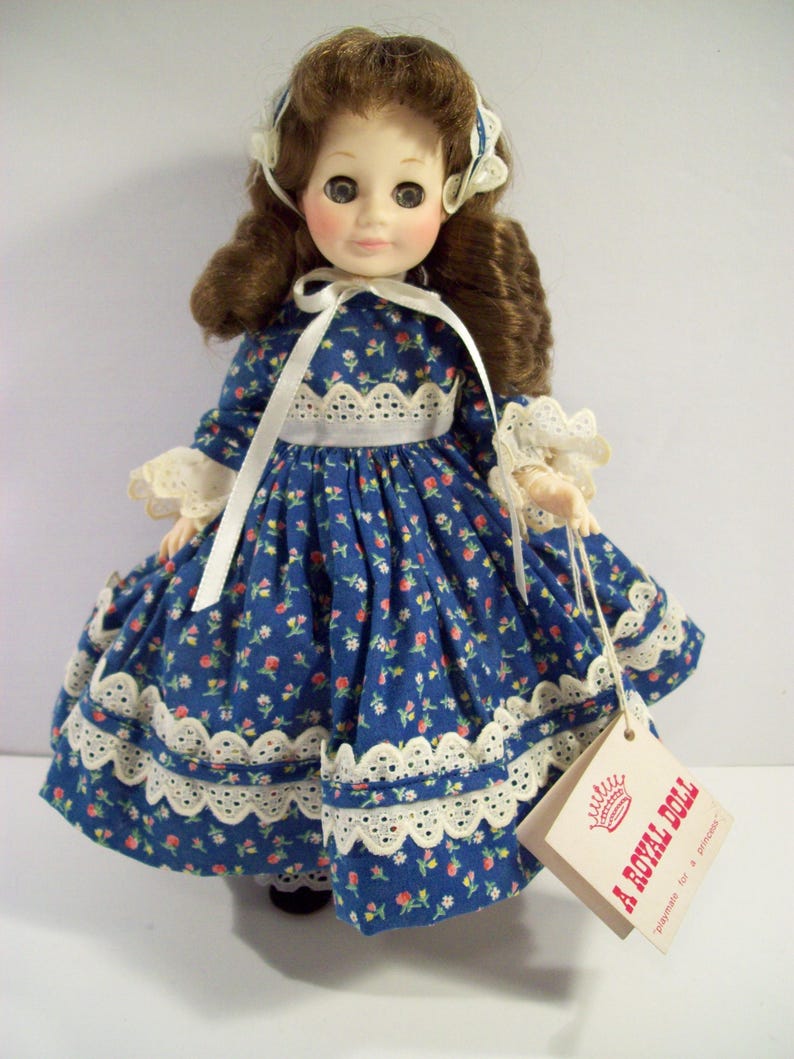 May include: A vintage porcelain doll wearing a blue and white floral dress with lace trim. The doll has brown hair and blue eyes. The doll is holding a tag that reads "A Royal Doll, Trademark for a product".