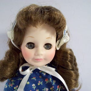 May include: A porcelain doll with brown hair styled in curls, wearing a blue and white floral dress with a white ribbon tied around the neck. The doll has blue eyes and rosy cheeks.