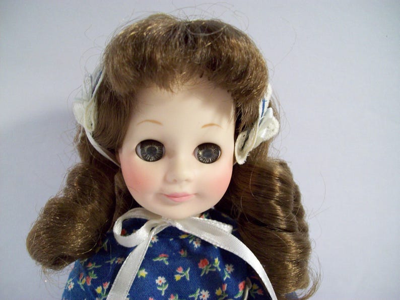 May include: A porcelain doll with brown hair styled in curls, wearing a blue and white floral dress with a white ribbon tied around the neck. The doll has blue eyes and rosy cheeks.