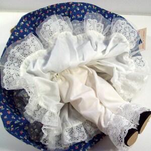 May include: A white doll with a blue and white floral print dress and white lace trim. The doll is wearing white underwear and has a white lace trim on the bottom of the dress.